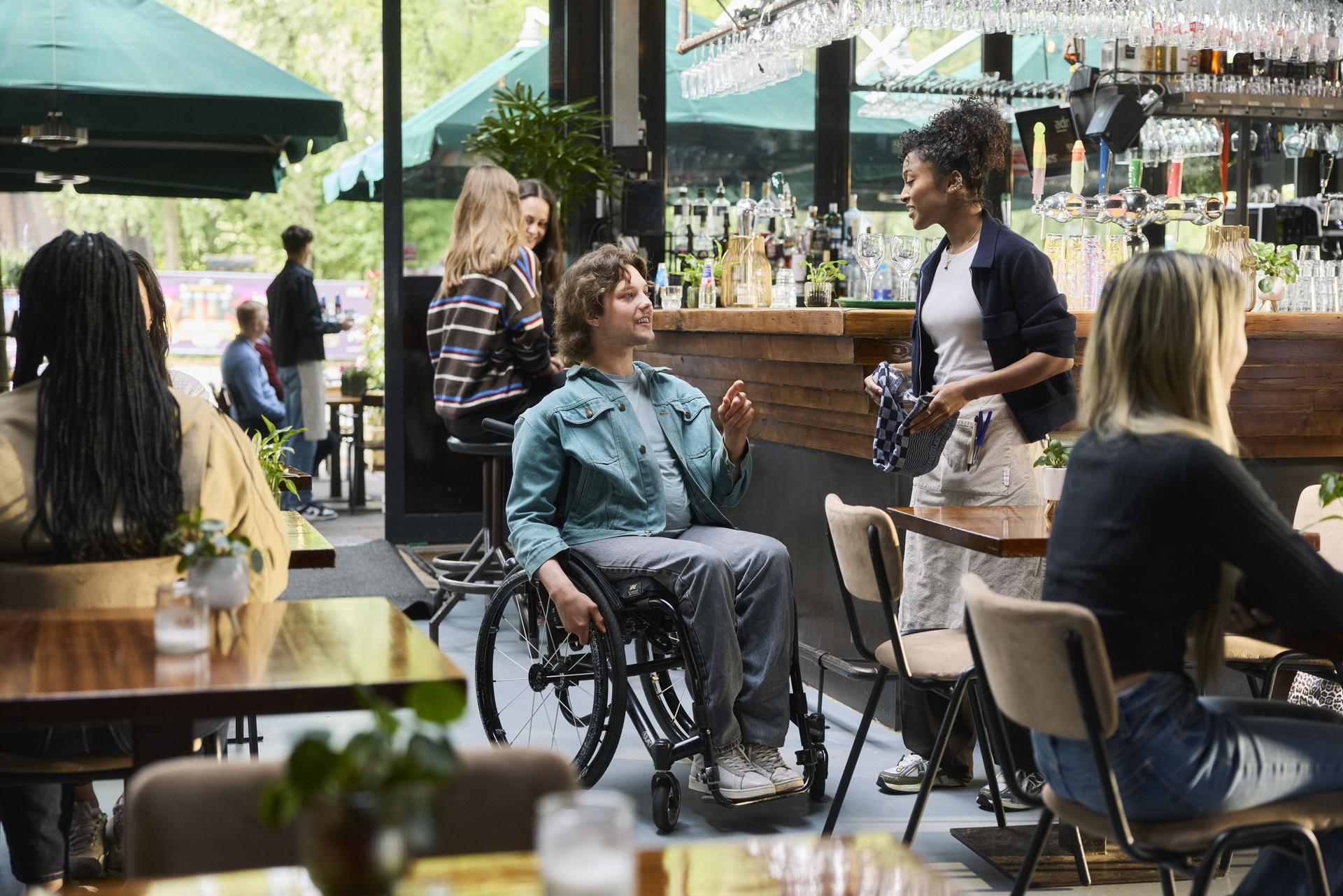 Campagnebeeld: Een man in een rolstoel zit in een café. Vrouwelijke ober is in gesprek met hem. 