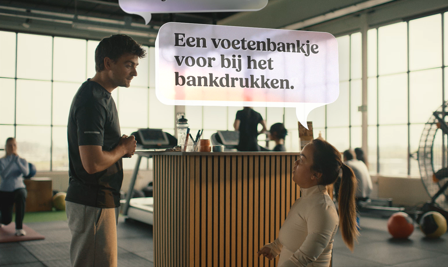 Still uit de campagnevideo ‘Heb je iets nodig?’ in een sportschool: een medewerker vraagt “Heb je iets nodig?”, waarop een vrouw antwoordt: “Een voetenbankje voor bij het bankdrukken.”