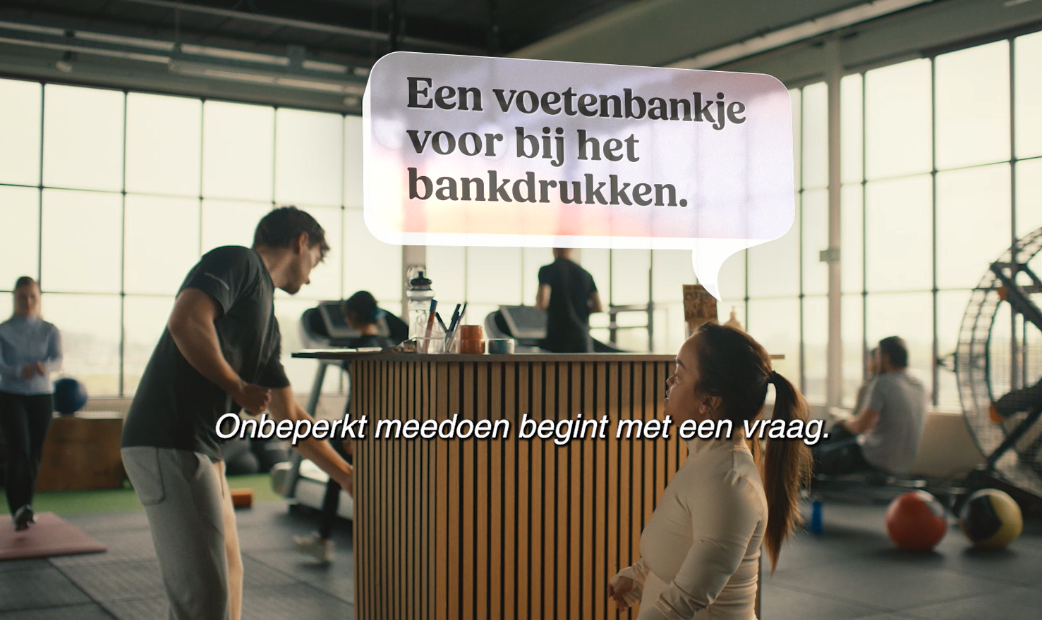 Still uit de campagnevideo ‘Heb je iets nodig?’ in een sportschool met ondertiteling: een medewerker vraagt “Heb je iets nodig?”, waarop een vrouw antwoordt: “Een voetenbankje voor bij het bankdrukken.”