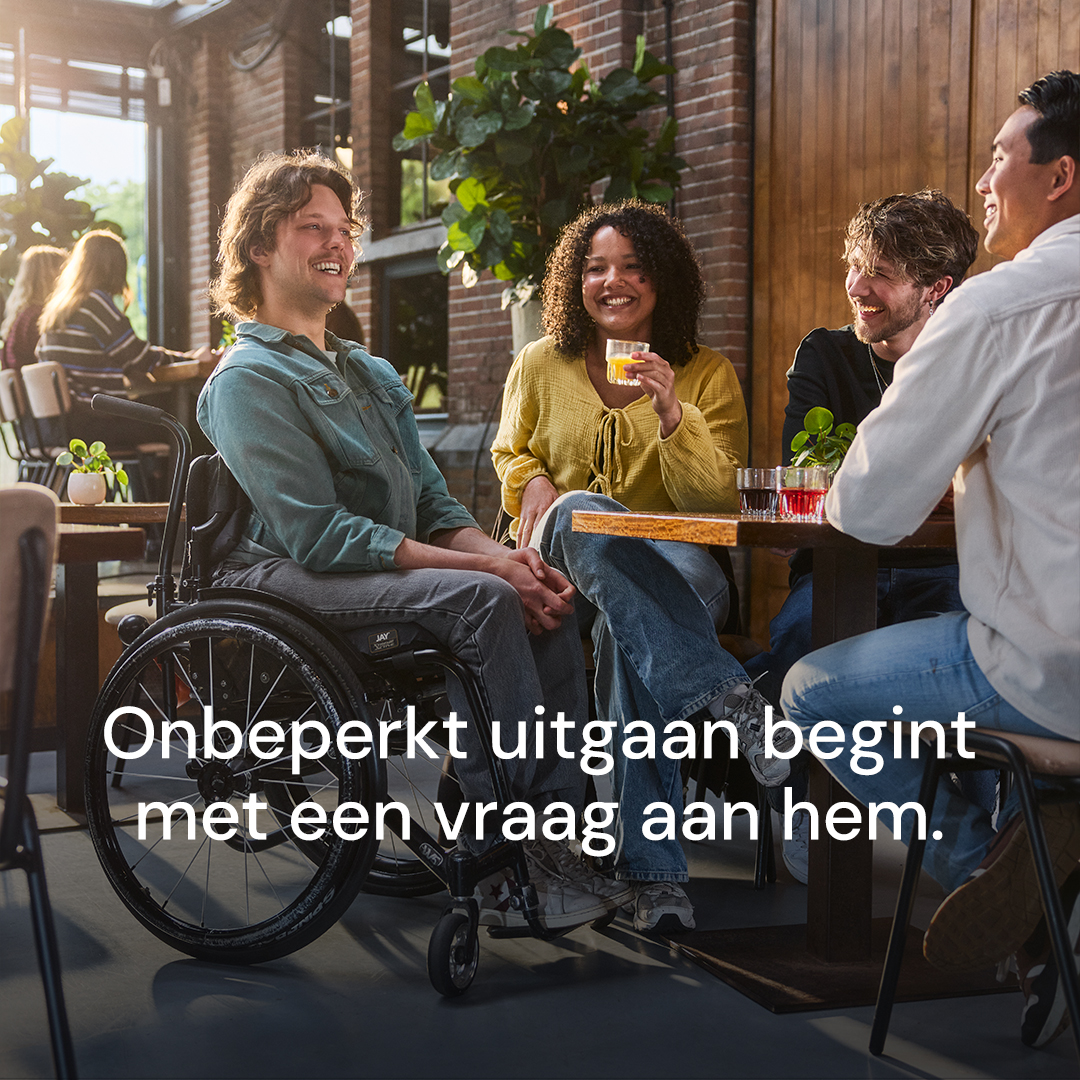 Campagnebeeld voor een carrousel: “Onbeperkt uitgaan begint met een vraag aan hem.” Een man in een rolstoel zit in een café aan tafel met drie anderen.