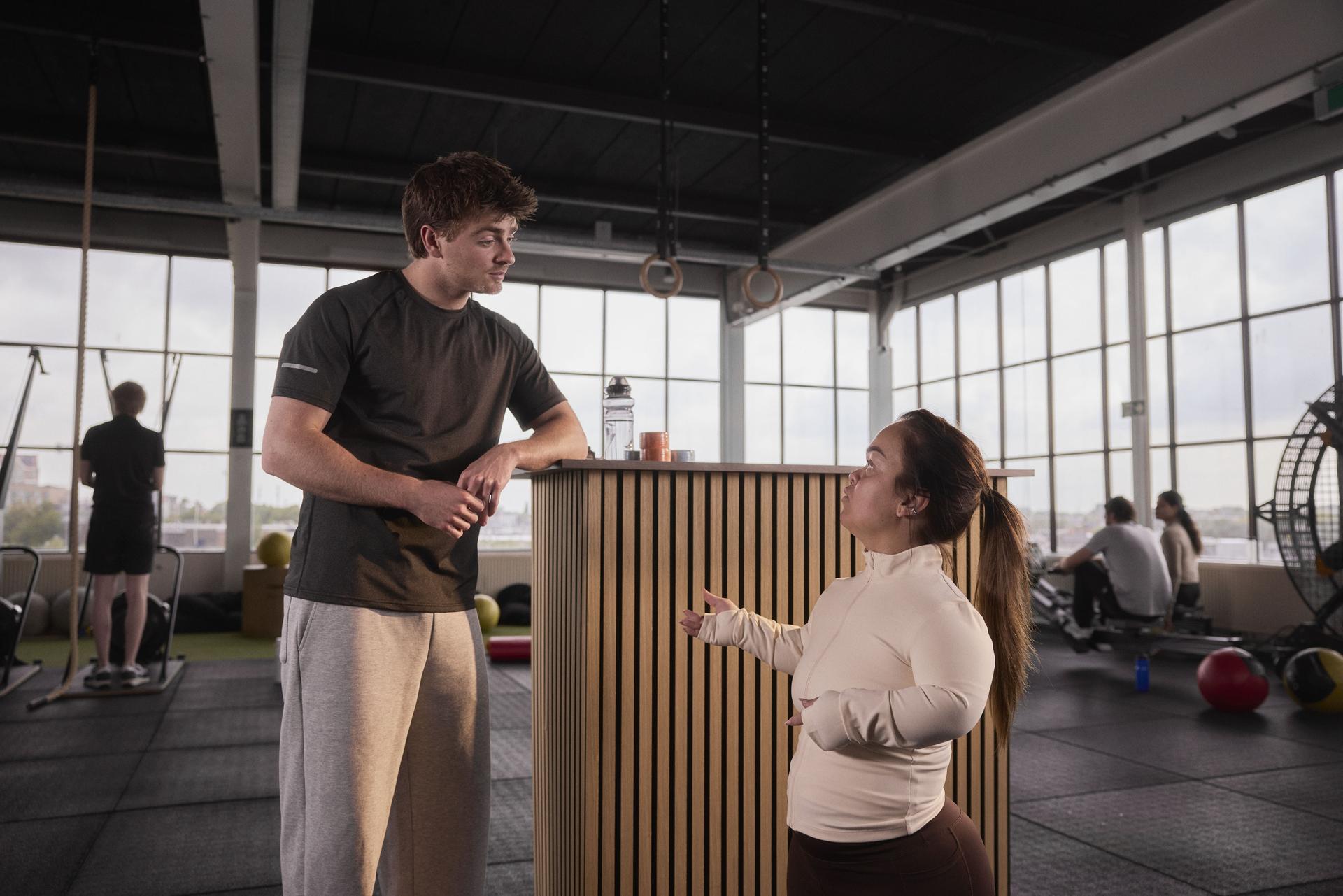 Campagnebeeld: Een vrouw (klein mens) is in gesprek met een man van de sportschool. 