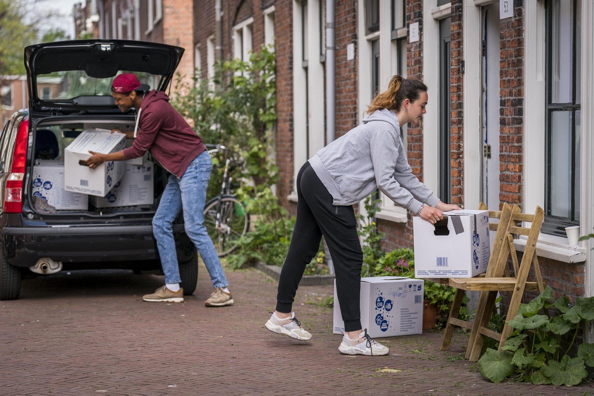 Twee mensen verhuizen dozen vanuit een auto naar een woning in een straat met rijtjeshuizen.